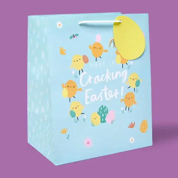 A light blue Easter gift bag decorated with cheerful cartoon chicks, eggs and flowers, with a yellow egg-shaped tag attached to the handle.